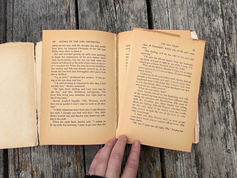 May include: An open vintage book with aged, cream-coloured paper pages featuring black printed text. The left page is titled Sandra of the Girl Orchestra and the right page is titled The Old Diary. The book shows signs of wear with visible binding.