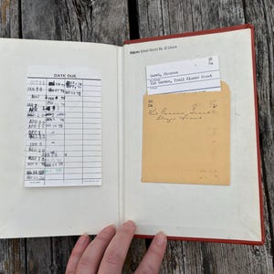 May include: An open hardcover book with a red spine, featuring a white library date due card with black ink stamps on the left page and a tan library pocket containing a white checkout card on the right page, marked with Kit Carson, Trail Blazer Scout.