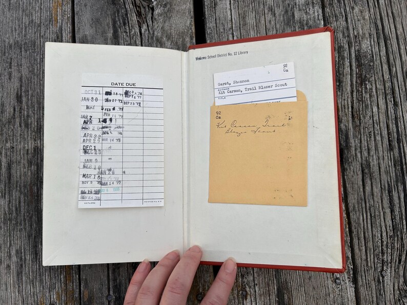 May include: An open hardcover book with a red spine, featuring a white library date due card with black ink stamps on the left page and a tan library pocket containing a white checkout card on the right page, marked with Kit Carson, Trail Blazer Scout.