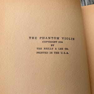 K&ouml;nnte beinhalten: Die Titelseite eines alten Buches mit dem Titel The Phantom Violin, Copyright 1934 von The Reilly & Lee Co., gedruckt in den U.S.A. Der Text ist in schwarzer Tinte auf gealtertem, hellbraunem Papier gedruckt, rechts ist ein Teilkapitelverzeichnis.