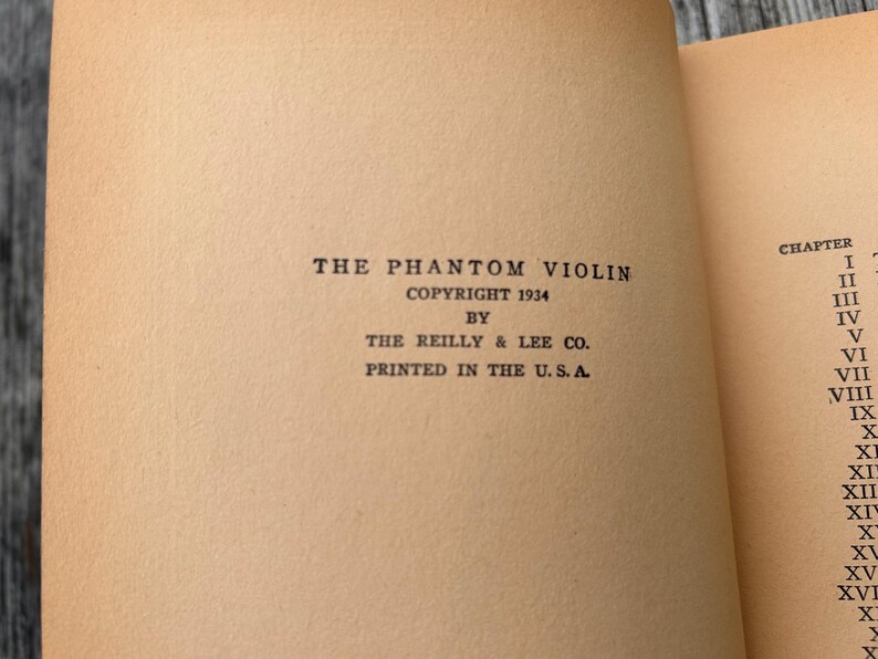 K&ouml;nnte beinhalten: Die Titelseite eines alten Buches mit dem Titel The Phantom Violin, Copyright 1934 von The Reilly & Lee Co., gedruckt in den U.S.A. Der Text ist in schwarzer Tinte auf gealtertem, hellbraunem Papier gedruckt, rechts ist ein Teilkapitelverzeichnis.
