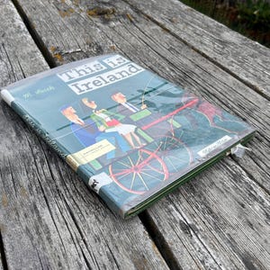 Peut inclure: Un livre reli&eacute; intitul&eacute; This is Ireland de M. Sasek, pr&eacute;sentant une illustration color&eacute;e de personnes dans une cal&egrave;che sur la couverture. Le livre est prot&eacute;g&eacute; par une couverture en plastique transparent et porte le logo de l'&eacute;diteur Macmillan.