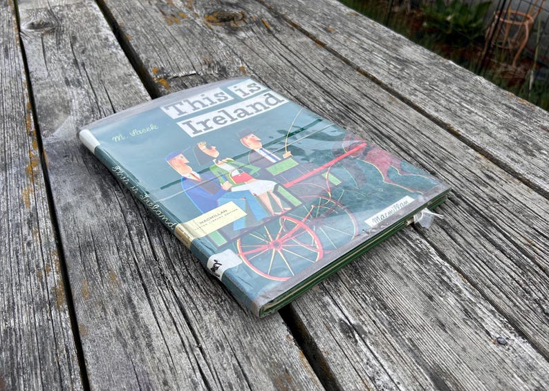 Peut inclure: Un livre reli&eacute; intitul&eacute; This is Ireland de M. Sasek, pr&eacute;sentant une illustration color&eacute;e de personnes dans une cal&egrave;che sur la couverture. Le livre est prot&eacute;g&eacute; par une couverture en plastique transparent et porte le logo de l'&eacute;diteur Macmillan.
