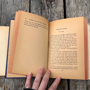 May include: An open vintage book with aged, tan-coloured pages showing printed black text. The left page displays the header Sandra of the Girl Orchestra, while the right page features Chapter Four titled The Test with several paragraphs of text.
