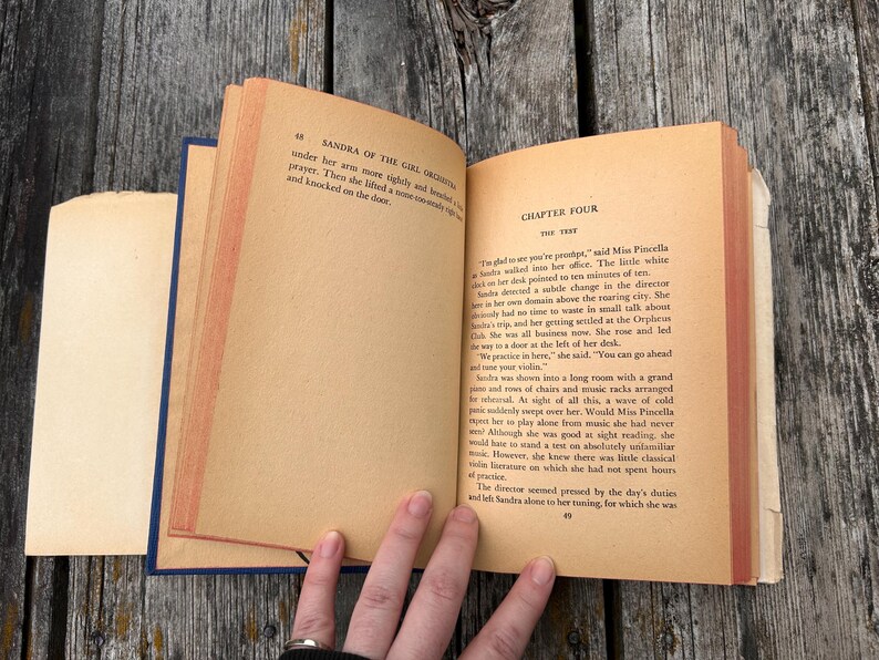 May include: An open vintage book with aged, tan-coloured pages showing printed black text. The left page displays the header Sandra of the Girl Orchestra, while the right page features Chapter Four titled The Test with several paragraphs of text.