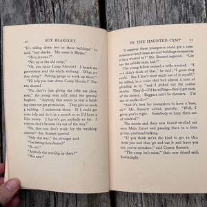 May include: An open book titled Roy Blakeley with pages 20 and 21 visible. The text includes dialogue about tearing down buildings at Camp Merritt. The paper is off-white with black printed text, featuring chapter headers and page numbers at the top.
