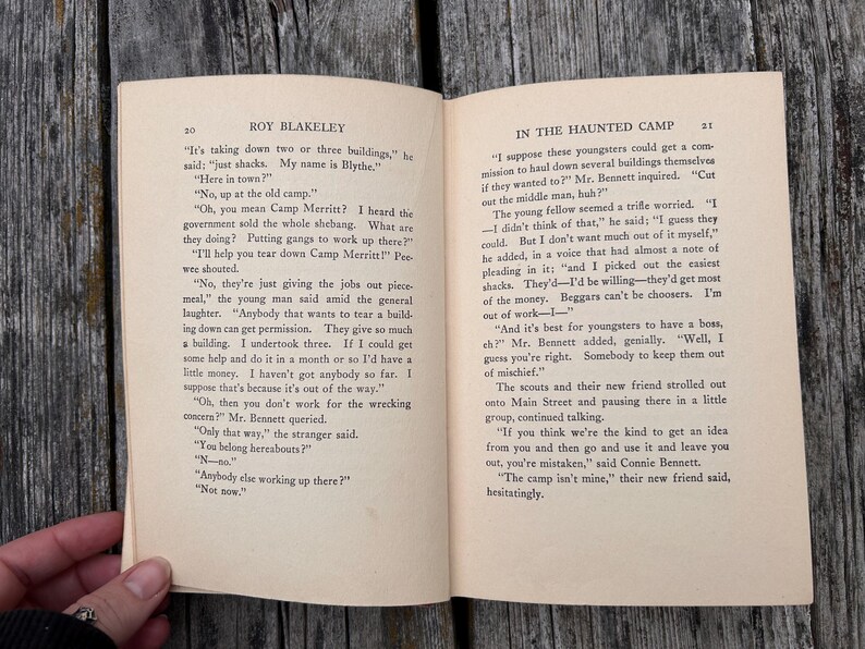 May include: An open book titled Roy Blakeley with pages 20 and 21 visible. The text includes dialogue about tearing down buildings at Camp Merritt. The paper is off-white with black printed text, featuring chapter headers and page numbers at the top.