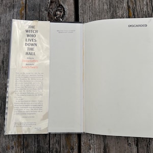 Puede incluir: Un libro de tapa dura abierto titulado The Witch Who Lives Down the Hall de Donna Guthrie e ilustrado por Amy Schwartz. La solapa interior delantera presenta un resumen impreso, mientras que la p&aacute;gina opuesta est&aacute; en blanco.