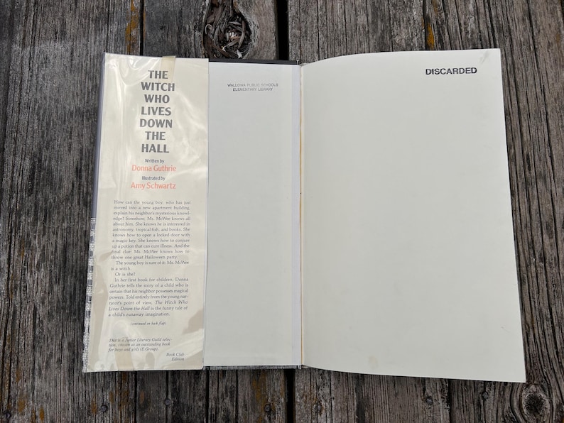 Puede incluir: Un libro de tapa dura abierto titulado The Witch Who Lives Down the Hall de Donna Guthrie e ilustrado por Amy Schwartz. La solapa interior delantera presenta un resumen impreso, mientras que la p&aacute;gina opuesta est&aacute; en blanco.