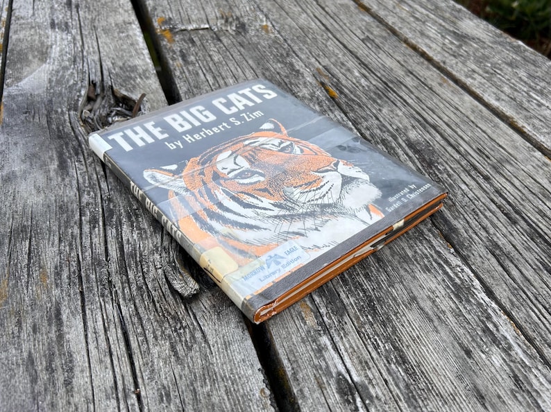 Puede incluir: Un libro de tapa dura titulado The Big Cats de Herbert S. Zim, con una ilustraci&oacute;n detallada de un tigre en la portada. El libro tiene una cubierta protectora de pl&aacute;stico de biblioteca e incluye el texto Library Edition y un logo de &aacute;guila.