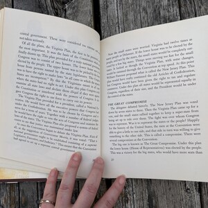 Puede incluir: Un libro abierto con p&aacute;ginas de color crema y texto impreso en negro. La p&aacute;gina izquierda trata sobre el Virginia Plan y la derecha detalla The Great Compromise. El libro se mantiene abierto, mostrando los n&uacute;meros de p&aacute;gina 26 y 27.