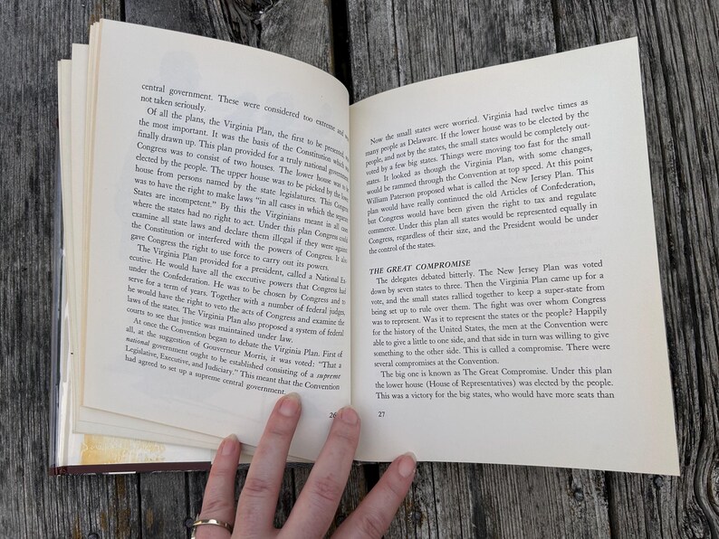Puede incluir: Un libro abierto con p&aacute;ginas de color crema y texto impreso en negro. La p&aacute;gina izquierda trata sobre el Virginia Plan y la derecha detalla The Great Compromise. El libro se mantiene abierto, mostrando los n&uacute;meros de p&aacute;gina 26 y 27.