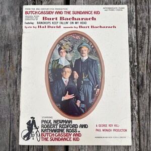 K&ouml;nnte beinhalten: Ein Vintage-Klaviernotenbuch f&uuml;r den Film Butch Cassidy and the Sundance Kid mit Musik von Burt Bacharach. Das Cover zeigt ein ovales Foto von drei Schauspielern in historischer Kleidung sowie Textkredite und eine Fahrradgrafik.