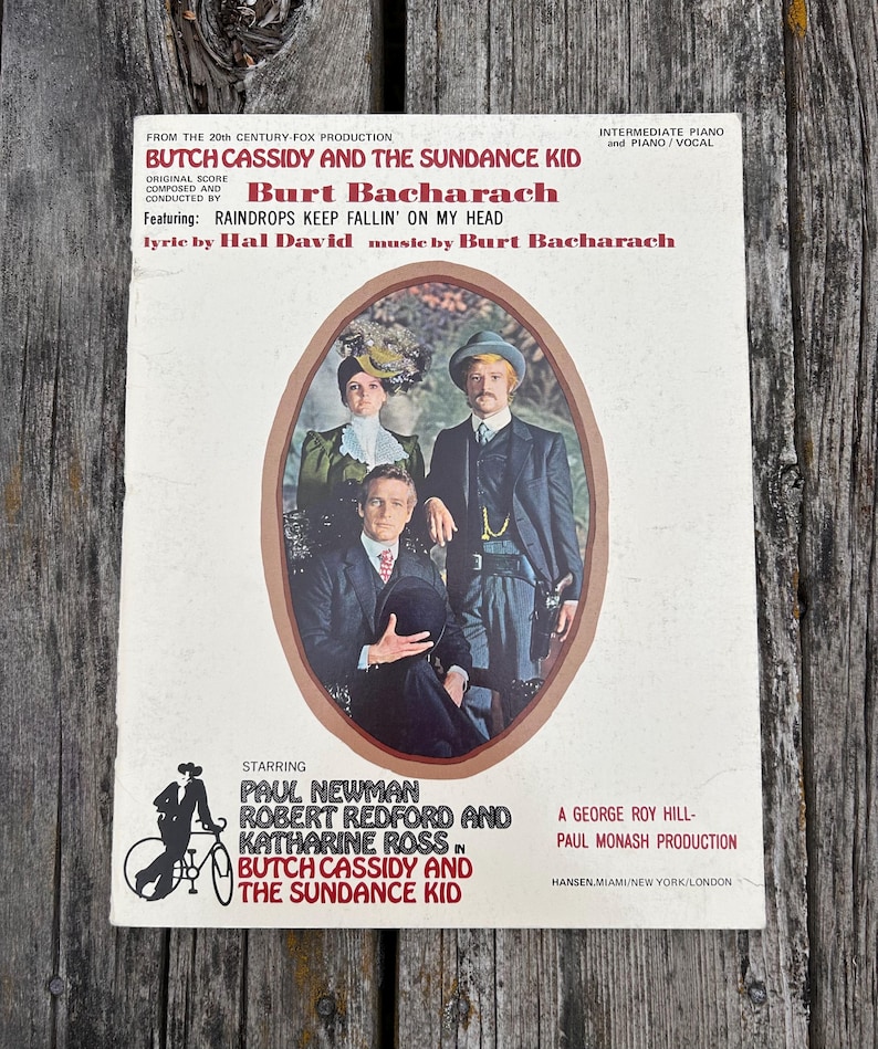 K&ouml;nnte beinhalten: Ein Vintage-Klaviernotenbuch f&uuml;r den Film Butch Cassidy and the Sundance Kid mit Musik von Burt Bacharach. Das Cover zeigt ein ovales Foto von drei Schauspielern in historischer Kleidung sowie Textkredite und eine Fahrradgrafik.