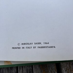 Peut inclure: Une page blanche de livre avec un texte imprim&eacute; en noir indiquant : &copy; MIROSLAV SASEK, 1964 PRINTED IN ITALY BY FABBRISTAMPA. La page pr&eacute;sente une bordure verte sur le bord inf&eacute;rieur et est plac&eacute;e sur une surface en bois.
