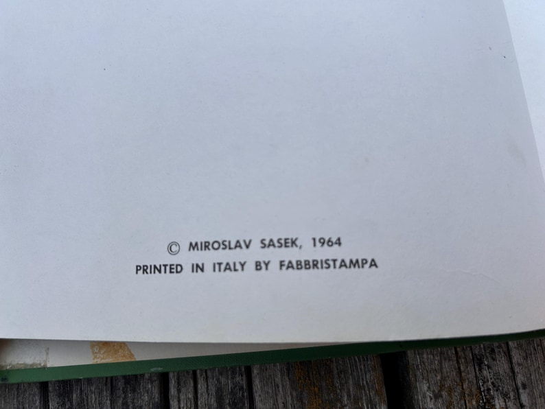 Peut inclure: Une page blanche de livre avec un texte imprim&eacute; en noir indiquant : &copy; MIROSLAV SASEK, 1964 PRINTED IN ITALY BY FABBRISTAMPA. La page pr&eacute;sente une bordure verte sur le bord inf&eacute;rieur et est plac&eacute;e sur une surface en bois.