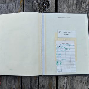 May include: An open book with cream-coloured pages. The right page features a vintage library checkout card with the text DATE DUE and various stamped dates in green ink, along with a smaller white card labelled with typed text and numbers.