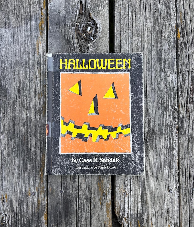 May include: A hardback book titled HALLOWEEN by Cass R. Sandak with illustrations by Frank Bozzo. The cover features a central orange jack-o'-lantern graphic with yellow triangular eyes and a jagged yellow mouth set against a black background.