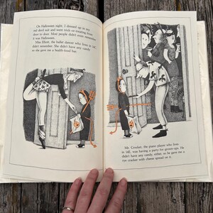 Puede incluir: Un libro infantil abierto con ilustraciones en blanco y negro que muestran a un ni&ntilde;o disfrazado de diablo pidiendo dulces. El texto describe encuentros de Halloween con vecinos, incluyendo a Miss Elliott y Mr. Crocket, con detalles naranjas.
