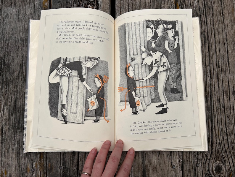 Puede incluir: Un libro infantil abierto con ilustraciones en blanco y negro que muestran a un ni&ntilde;o disfrazado de diablo pidiendo dulces. El texto describe encuentros de Halloween con vecinos, incluyendo a Miss Elliott y Mr. Crocket, con detalles naranjas.
