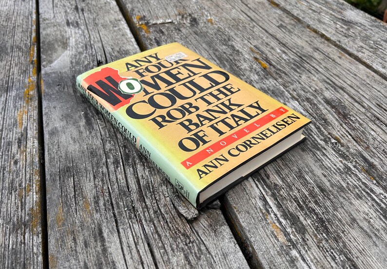 May include: A hardcover book titled Any Four Women Could Rob the Bank of Italy by Ann Cornelisen. The cover features a yellow to white gradient background with bold black text, a red accent bar, and a small green circular graphic on the front.