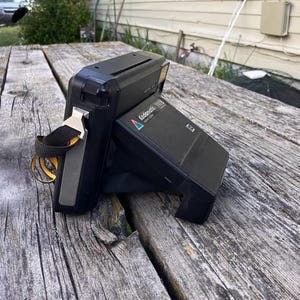 Peut inclure: Un appareil photo instantané Kodak Kodamatic 980L noir avec une sangle noire et un fermoir en métal argenté. L'appareil photo est posé sur une surface en bois vieilli. Le logo Kodamatic est visible sur le côté.