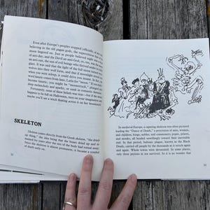 以下が含まれることがあります： 白いページに黒い文字が印刷された開いた本。左ページにはSKELETONという見出しと説明文があり、右ページには王冠をかぶった人物が人々の集団と骸骨を先導する様子を描いた黒と白のインク画が掲載されています。