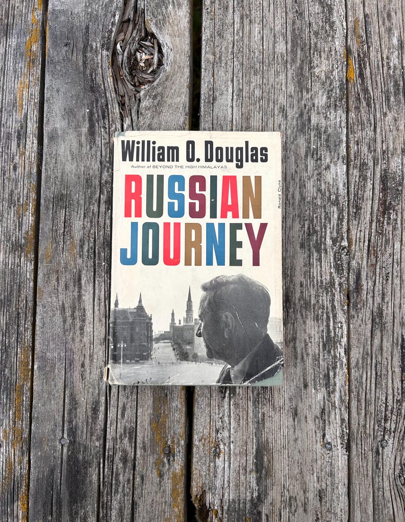 May include: A hardback book titled Russian Journey by William O. Douglas. The cover features a black and white photograph of a man in profile overlooking a city square with tall buildings, and the title text in red, blue, and brown.