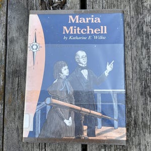 Puede incluir: Un libro de tapa dura titulado Maria Mitchell de Katharine E. Wilkie, con una ilustraci&oacute;n de portada azul de una persona se&ntilde;alando al cielo nocturno junto a un telescopio, con una rosa de los vientos a la izquierda y el texto A Discovery Book.