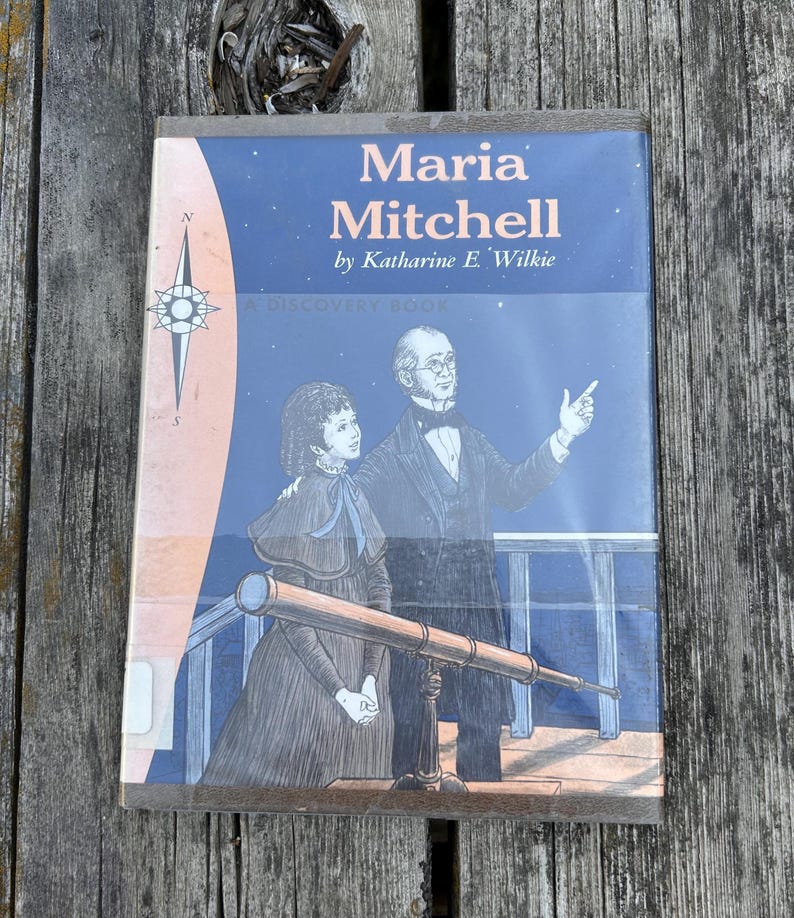 Puede incluir: Un libro de tapa dura titulado Maria Mitchell de Katharine E. Wilkie, con una ilustraci&oacute;n de portada azul de una persona se&ntilde;alando al cielo nocturno junto a un telescopio, con una rosa de los vientos a la izquierda y el texto A Discovery Book.