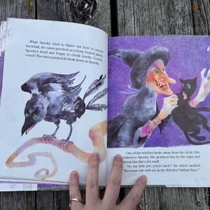 K&ouml;nnte beinhalten: Ein aufgeschlagenes Kinderbuch mit farbenfrohen Illustrationen eines schwarzen Raben auf einem Ast und einer Hexe, die eine schwarze Katze h&auml;lt. Die Seiten enthalten gedruckten Text, der die Szene beschreibt, mit violetten und wei&szlig;en Hintergr&uuml;nden.