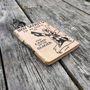 K&ouml;nnte beinhalten: Ein gebundenes Vintage-Buch mit dem Titel Boy Scout's in Alaska or The Camp on the Glacier. Der Einband ist hellbraun mit schwarzem Text und einer schwarzen Strichzeichnung eines Pfadfinders mit Flagge. Das Buch weist Gebrauchsspuren auf.