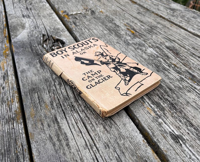 K&ouml;nnte beinhalten: Ein gebundenes Vintage-Buch mit dem Titel Boy Scout's in Alaska or The Camp on the Glacier. Der Einband ist hellbraun mit schwarzem Text und einer schwarzen Strichzeichnung eines Pfadfinders mit Flagge. Das Buch weist Gebrauchsspuren auf.