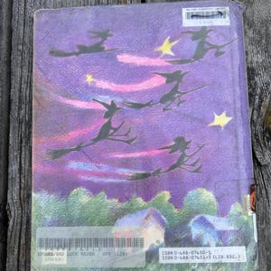 K&ouml;nnte beinhalten: Ein rechteckiges gebundenes Buch mit einer Illustration eines violetten Nachthimmels, die Silhouetten von vier auf Besen fliegenden Hexen, gelbe Sterne und rosa Streifen zeigt. Unten sind gr&uuml;ne B&auml;ume und zwei kleine H&auml;user zu sehen.