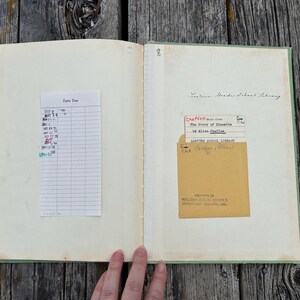 K&ouml;nnte beinhalten: Ein aufgeschlagenes altes Buch mit hellgr&uuml;nem Einband, das auf der linken Seite einen wei&szlig;en Leihzettel und auf der rechten Seite eine beige Bibliothekstasche mit Karte enth&auml;lt. Der Text umfasst Lostine Grade School Library und The Story of Nimble.