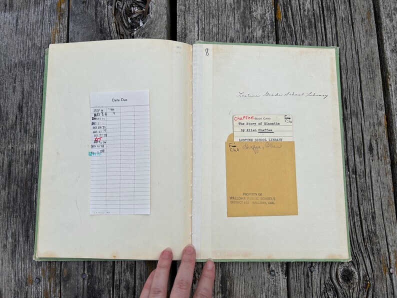 K&ouml;nnte beinhalten: Ein aufgeschlagenes altes Buch mit hellgr&uuml;nem Einband, das auf der linken Seite einen wei&szlig;en Leihzettel und auf der rechten Seite eine beige Bibliothekstasche mit Karte enth&auml;lt. Der Text umfasst Lostine Grade School Library und The Story of Nimble.