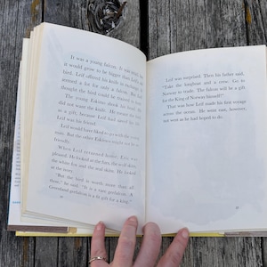 Pu&ograve; includere: Un libro con copertina rigida aperto con pagine color crema e testo stampato in nero. La pagina sinistra contiene paragrafi su un giovane falco e una persona, mentre la pagina destra continua la narrazione su Leif e un viaggio in Norvegia.
