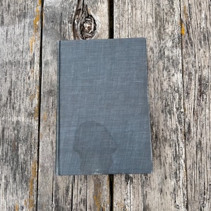 May include: A rectangular hardcover book with a dark gray, textured fabric cover. The front cover features a subtle, darker circular stain or mark near the bottom center, and the book is positioned vertically on a weathered wooden surface.
