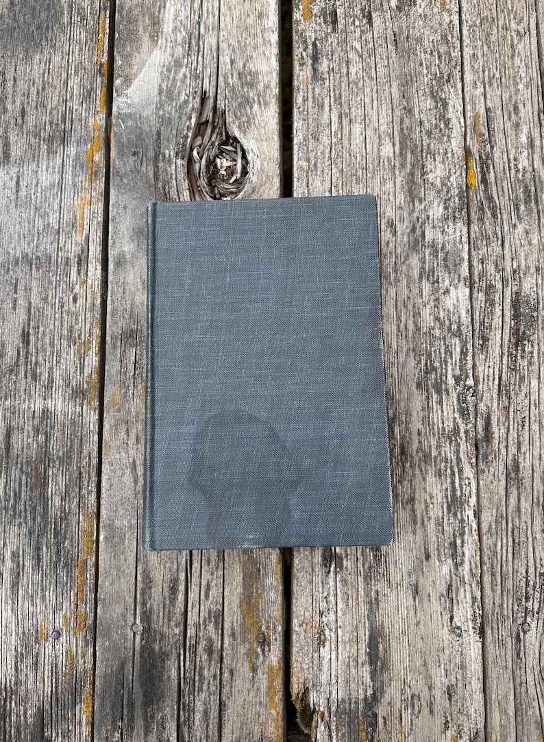May include: A rectangular hardcover book with a dark gray, textured fabric cover. The front cover features a subtle, darker circular stain or mark near the bottom center, and the book is positioned vertically on a weathered wooden surface.