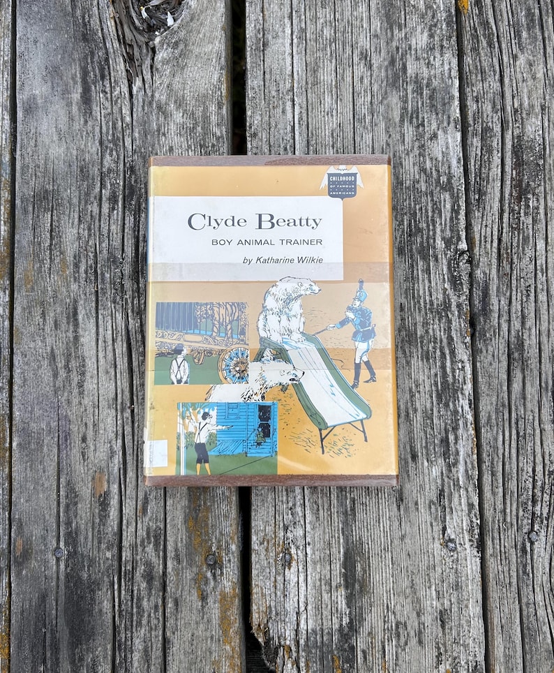 May include: A hardback book titled Clyde Beatty Boy Animal Trainer by Katharine Wilkie. The tan cover features illustrations of a polar bear on a slide, a trainer, and a person standing near a blue building. A small badge reads Childhood of Famous Americans.