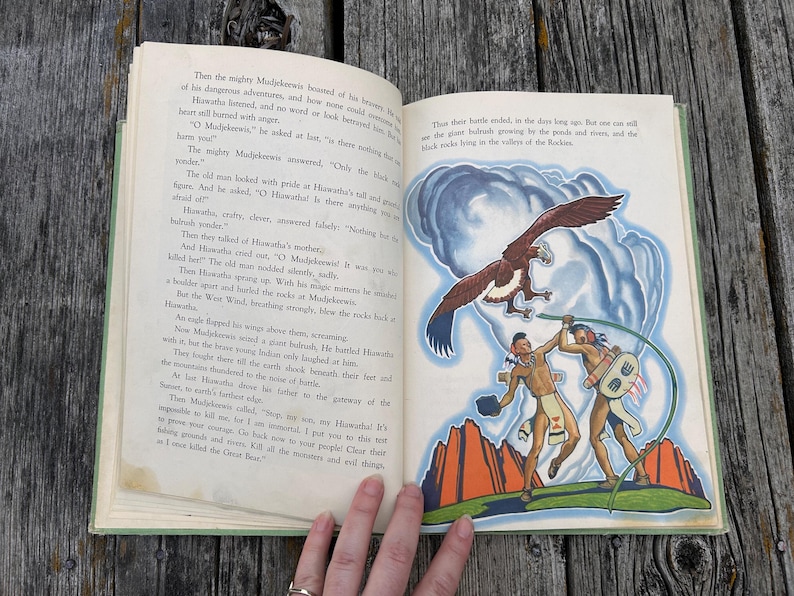 K&ouml;nnte beinhalten: Ein aufgeschlagenes altes Buch mit gedrucktem Text auf der linken Seite und einer farbigen Illustration auf der rechten Seite. Das Bild zeigt zwei Figuren in traditioneller Kleidung, einen gro&szlig;en braunen Adler und rote Felsen vor blauem Himmel.