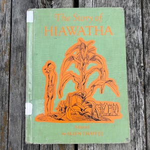 K&ouml;nnte beinhalten: Ein hellgr&uuml;nes gebundenes Buch mit dem Titel The Story of HIAWATHA, adaptiert von ALLEN CHAFFEE. Das Cover zeigt eine orangefarbene grafische Illustration einer Person neben einer gro&szlig;en Maispflanze, mit kleineren Figuren im Hintergrund.