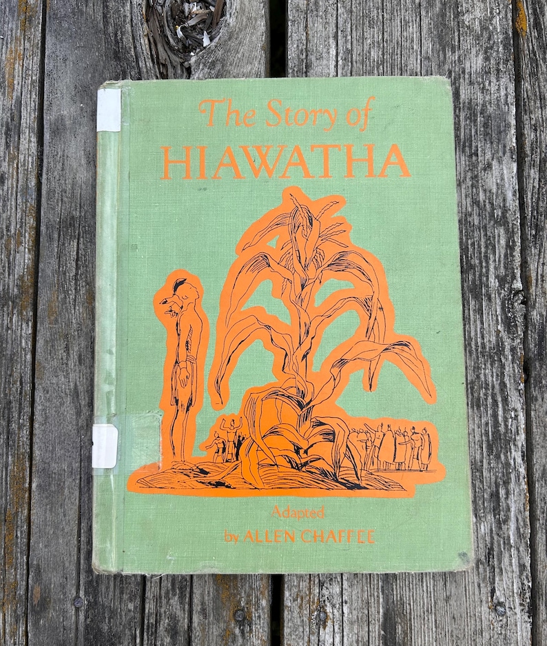 K&ouml;nnte beinhalten: Ein hellgr&uuml;nes gebundenes Buch mit dem Titel The Story of HIAWATHA, adaptiert von ALLEN CHAFFEE. Das Cover zeigt eine orangefarbene grafische Illustration einer Person neben einer gro&szlig;en Maispflanze, mit kleineren Figuren im Hintergrund.