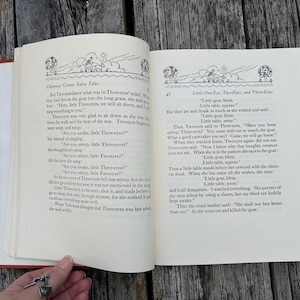 May include: An open hardcover book with cream-colored pages featuring black printed text and illustrations. The left page is titled Chimney Corner Fairy Tales, and the right page is titled Little One-Eye, Two-Eyes, and Three-Eyes with story text.