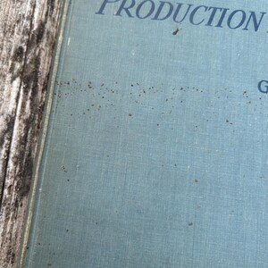Pu&ograve; includere: Una copertina rigida di colore azzurro e testurizzata con testo in blu scuro che recita "PRODUCTION" e "G". La superficie della copertina mostra segni visibili di invecchiamento, tra cui macchie marrone scuro sparse e una consistenza simile al tessuto.