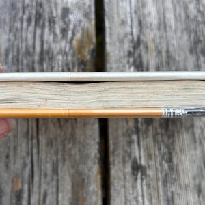 May include: A thin, rectangular book with a white spine and off-white pages, positioned horizontally alongside a yellow wooden pencil with a silver metal ferrule and a small, light blue eraser tip, showing the side profile of the book.