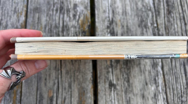 May include: A thin, rectangular book with a white spine and off-white pages, positioned horizontally alongside a yellow wooden pencil with a silver metal ferrule and a small, light blue eraser tip, showing the side profile of the book.