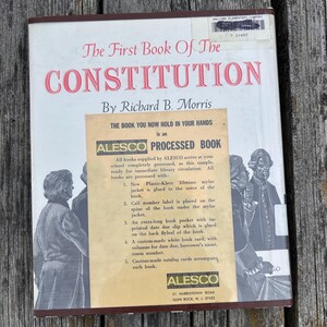 Puede incluir: Un libro de tapa dura titulado The First Book Of The Constitution de Richard B. Morris. La cubierta blanca presenta ilustraciones en blanco y negro de figuras hist&oacute;ricas y una pegatina beige de Alesco.