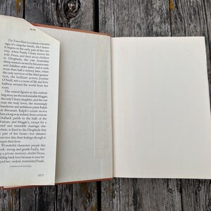 May include: An open hardcover book with a cream-colored dust jacket flap on the left side. The flap contains printed text describing the plot of the novel The Thorn Birds, including characters like Paddy Cleary, Fiona, Meggie, and Ralph de Bricassart.