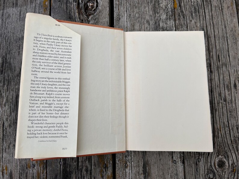 May include: An open hardcover book with a cream-colored dust jacket flap on the left side. The flap contains printed text describing the plot of the novel The Thorn Birds, including characters like Paddy Cleary, Fiona, Meggie, and Ralph de Bricassart.